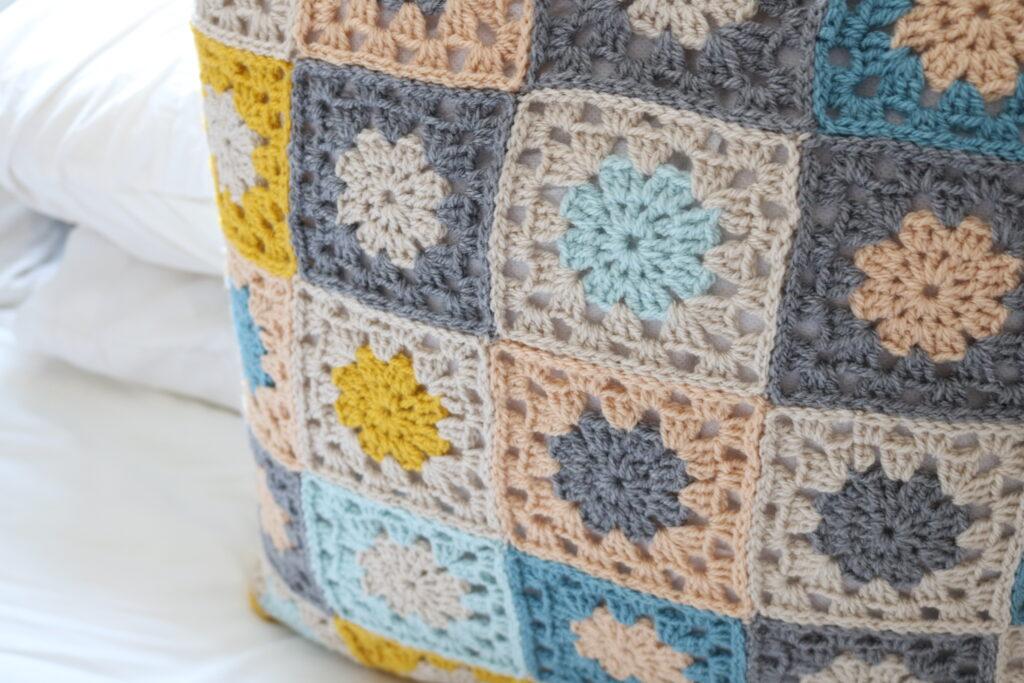 A cushion made from granny squares sits on a white bed. The cushion is made from brown, grey, blue and mustard yarn.