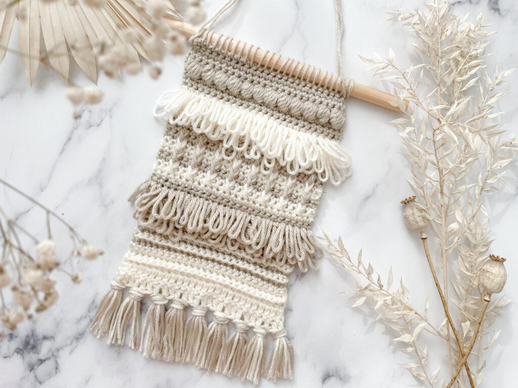 a flat lay image of a textured crochet wall hanging in colours cream and beige on a marbled back drop surrounds by dried flowers.