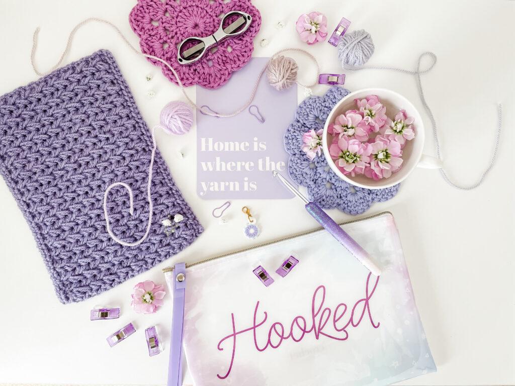 flay lay image of purple objects including a purple crochet swatch of the feather stitch, coaster, purple flowers, stitch markers and a pencil case that says hooked on it.