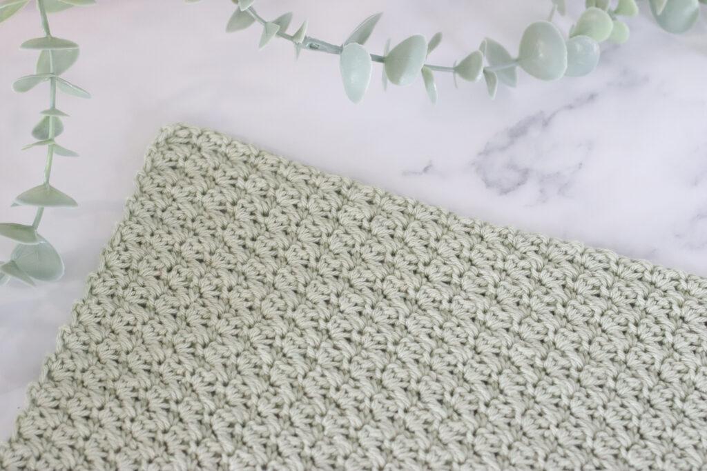 a pale green crochet swatch of the suzette stitch on a marbled backdrop and foliage to the left of the image.
