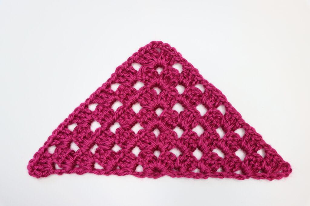 A granny square triangle made from berry coloured yarn sits on a white background.