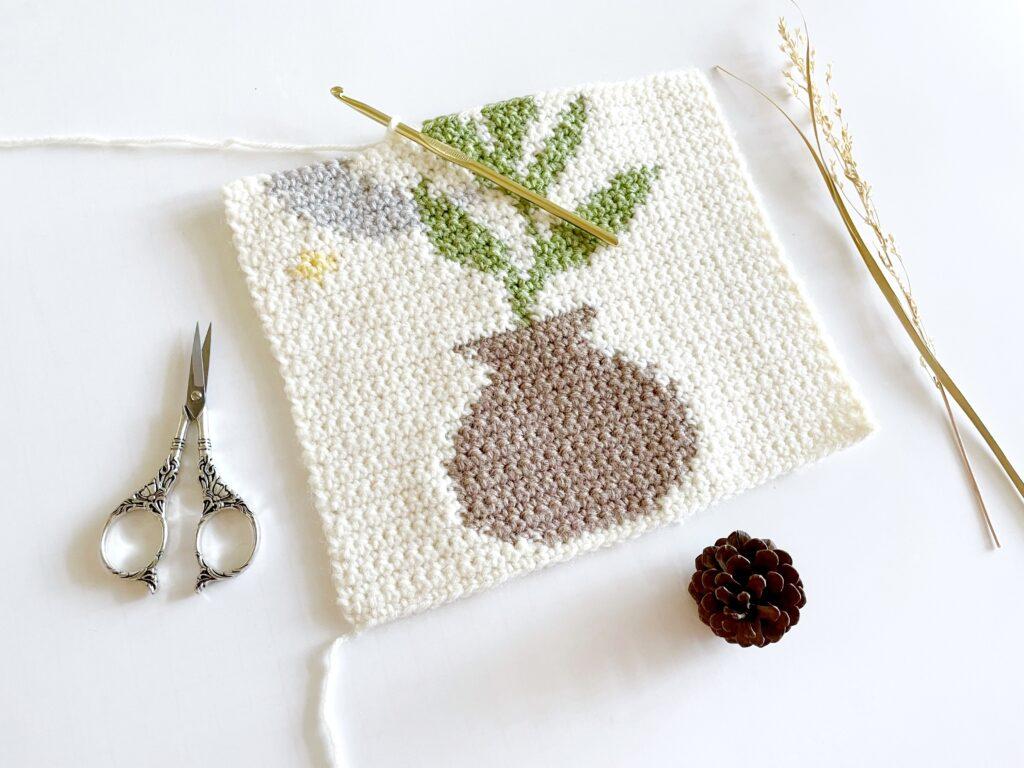 The crochet wall hanging is in progress and is displayed besides a pair of silver scissors and a pinecone. 