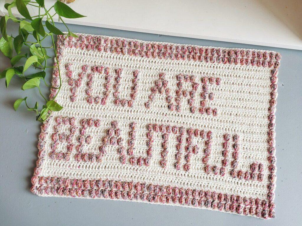 a flat lay image of a textured crochet bath mat on the floor that says You are beautiful within the stitches.