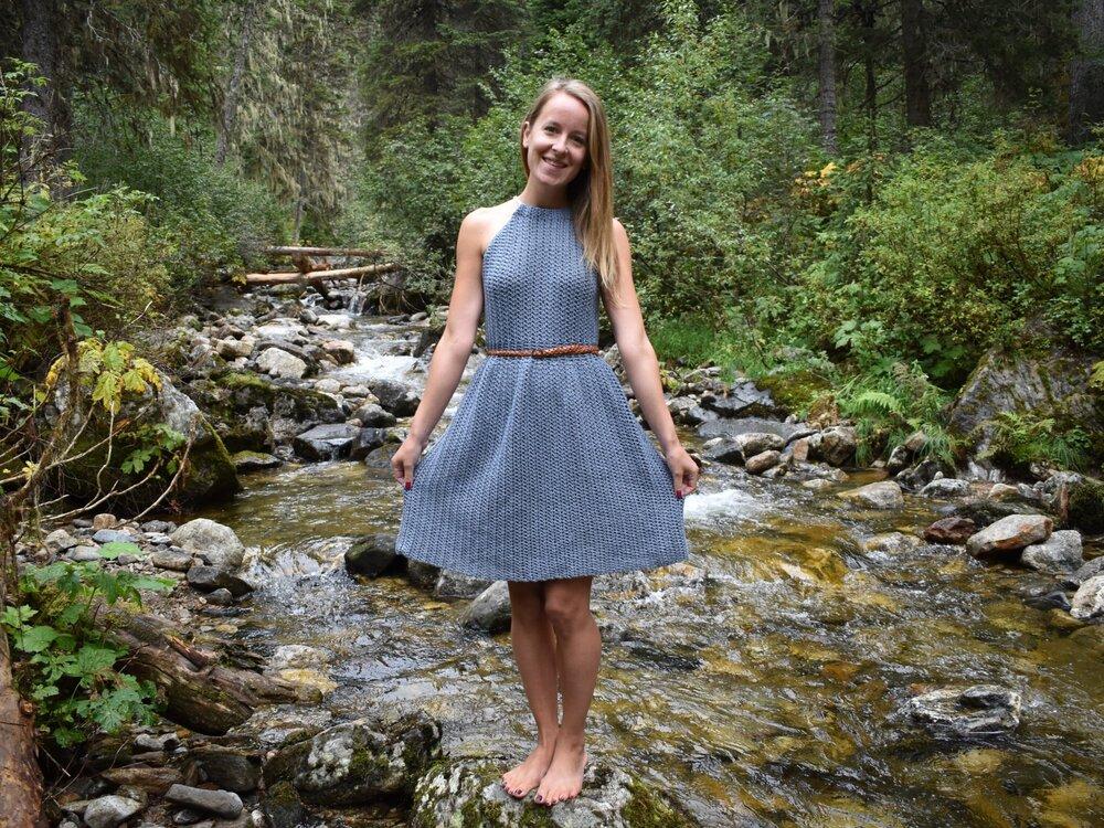 A woman stands in a stream wearing a blue halterneck crochet dress. It is cinched in with a brown belt. She is barefoot and smiling.