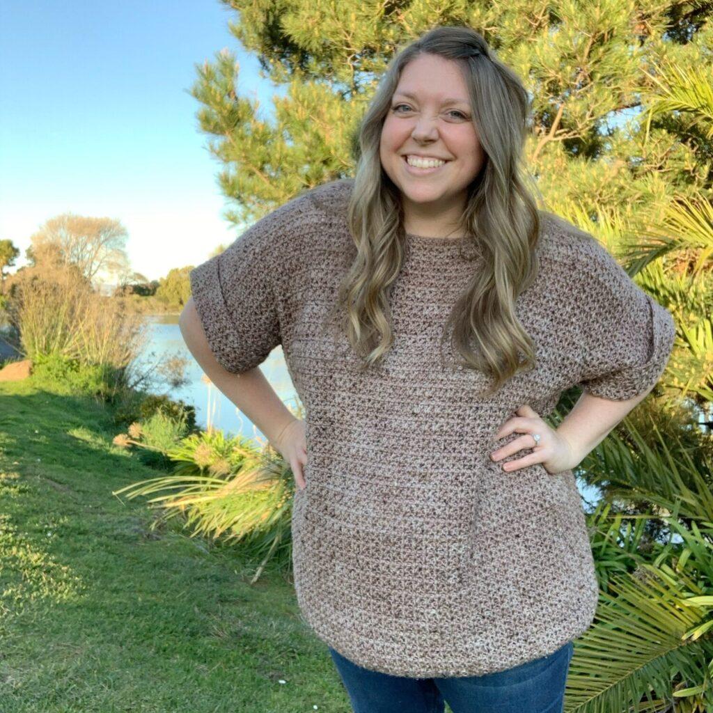 A woman stands surrounded by trees, she is wearing a taupe coloured crochet tee in a slouchy style. She is smiling.