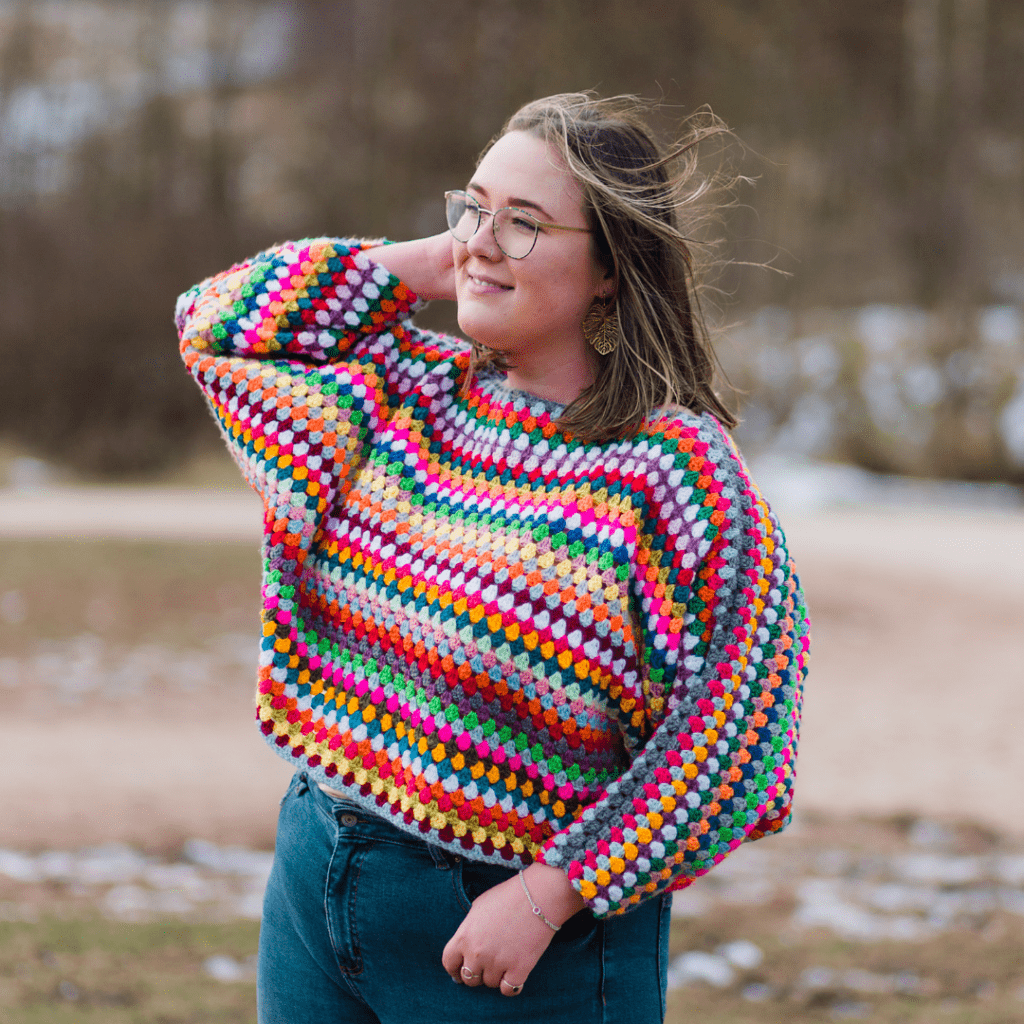 A woman looks to the left, she is wearing a rainbow-coloured granny stripe sweater. She is smiling.