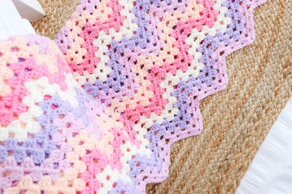 A granny ripple blanket in shades of lilac, purple, pink and cream is laid over a white miniature bed with a straw mat directly under the blanket.