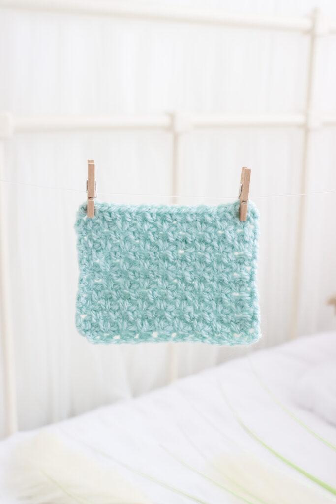 A swatch of moss stitch aka linen stitch in aqua yarn is pegged to a line with small wooden clothes pegs. The background is completely white.