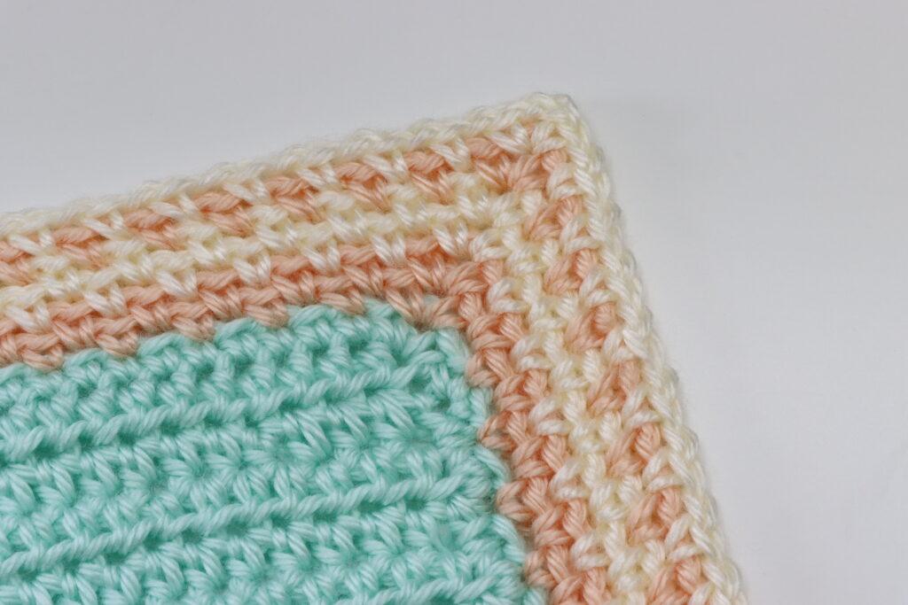 A swatch of crochet sits on a white background. The moss stitch border is displayed around a plain crochet swatch in aqua yarn. The border is striped in peach and cream.