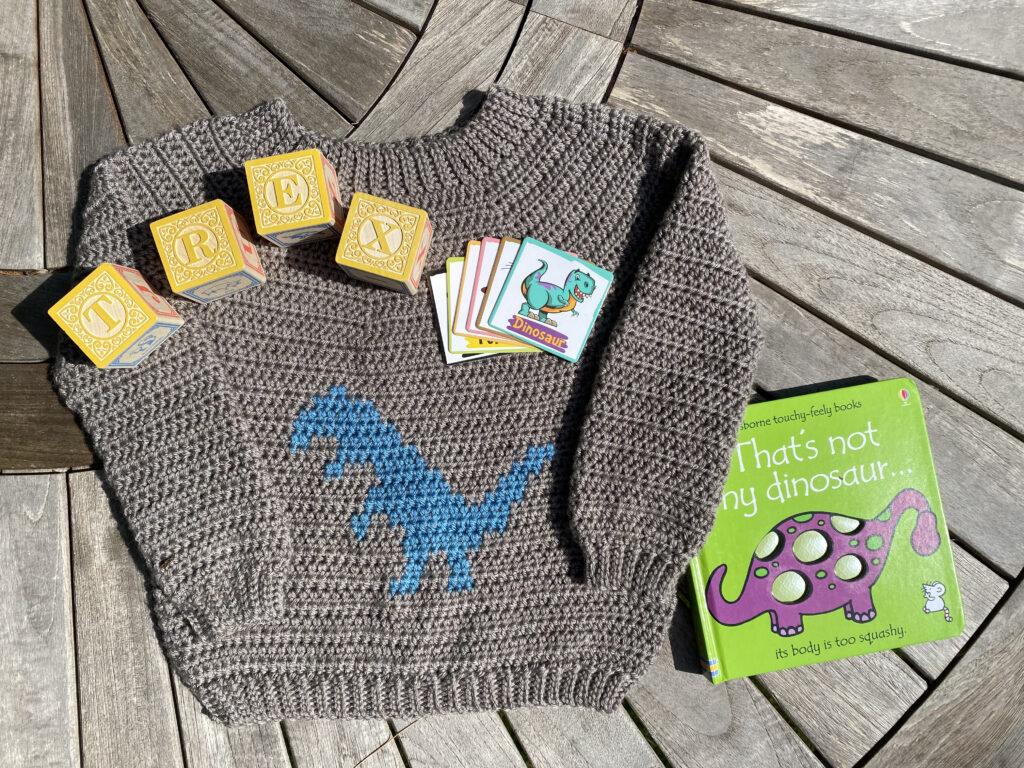 A crochet sweater made of brown yarn sits on a wooden board. The sweater has a blue dinosaur motif on the front. Surrounding the sweater are dino flash cards, a dinosaur book and the Premier Anti Pilling DK yarn used to make the sweater.