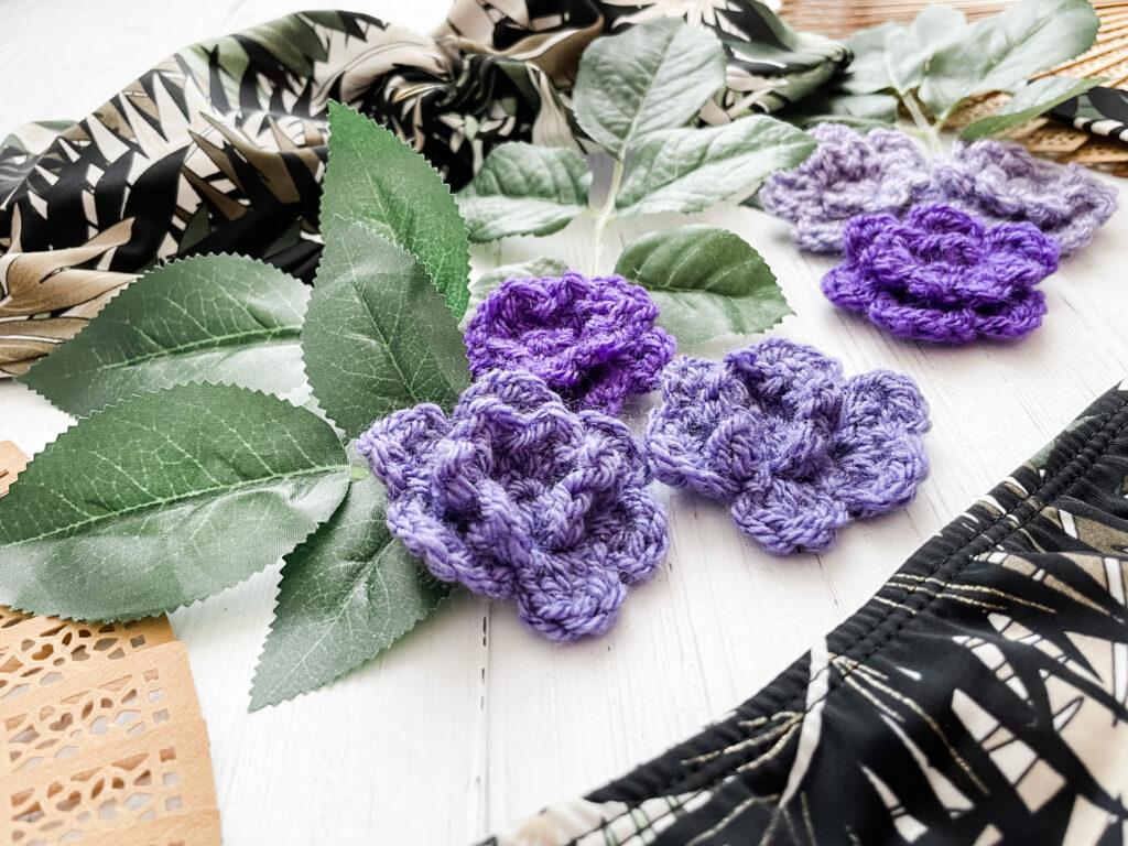 A collection of purple crochet flowers in different tones of the colour sit on a white wooden background. Beside them is an open wooden fan and a leaf print bikini in white and deep forest green.