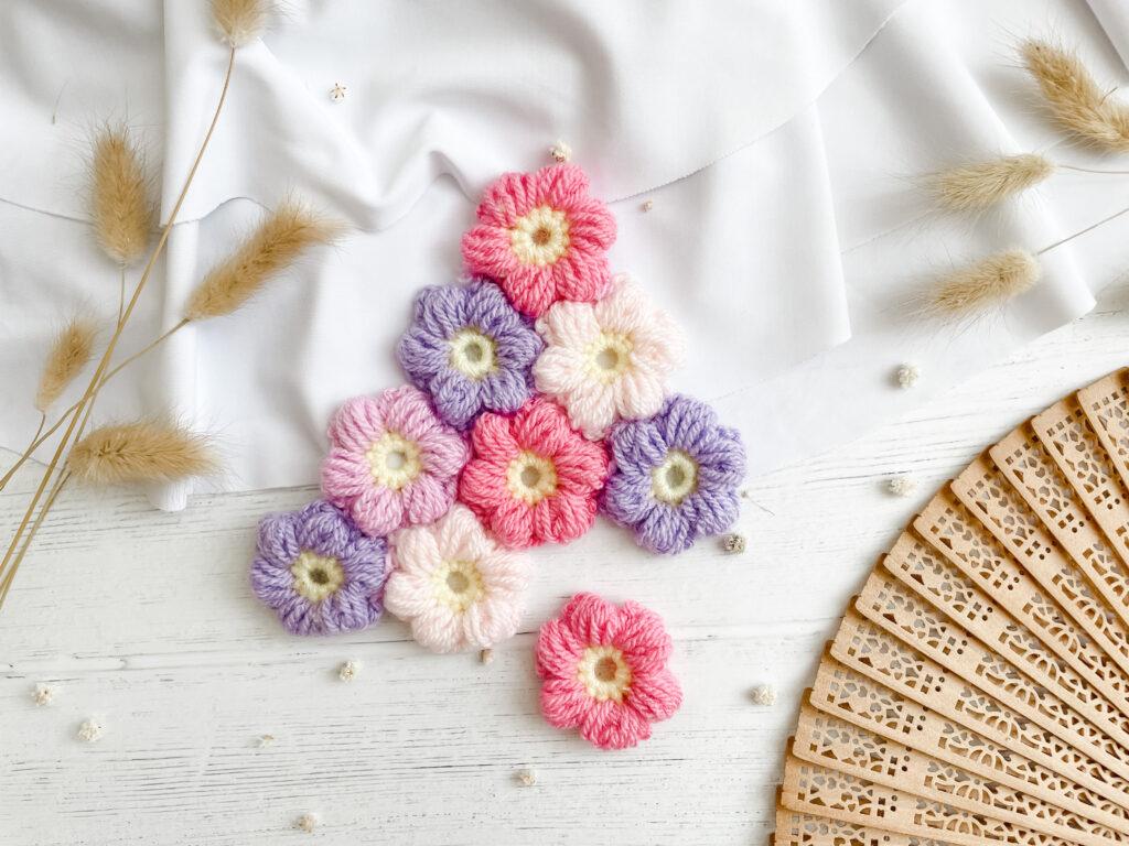 A collection of crochet puff flowers in pinks, purples and cream, all with yellow centres sit on a white wooden background beside an open wooden fan.