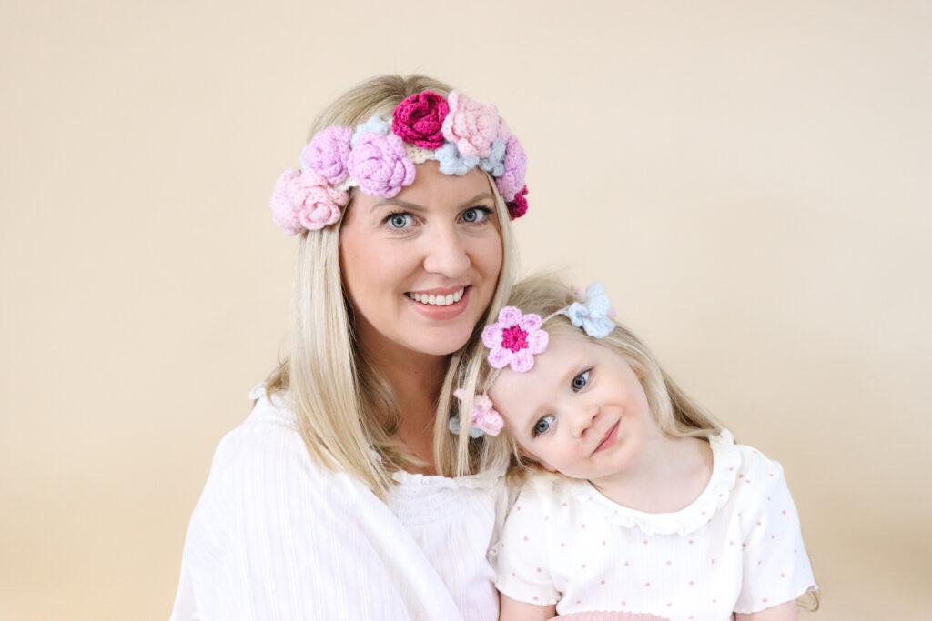 an image of mother and daughter sat infront of a cream backdrop wearing a crochet flower crown each made in pink, purple and pale blue yarn.