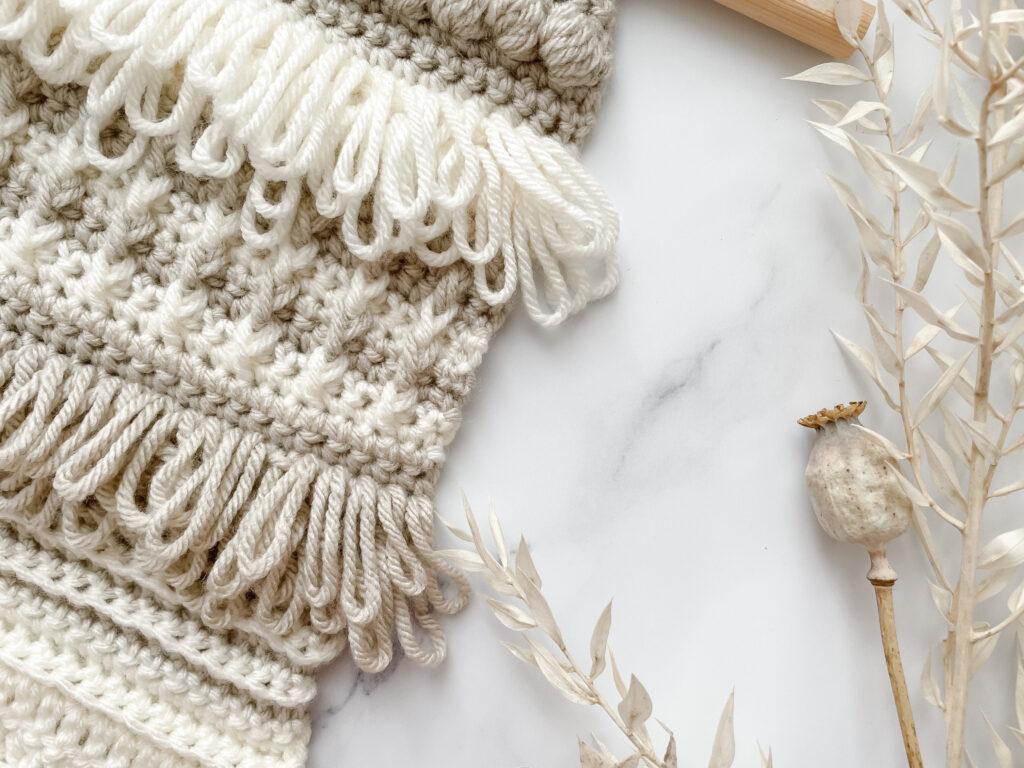 A crochet wall hanging made in cream and light brown yarn sits on a white marble background. The wall hanging is made up of different stitches. Shown here prominently are loop stitch and bar stitch.