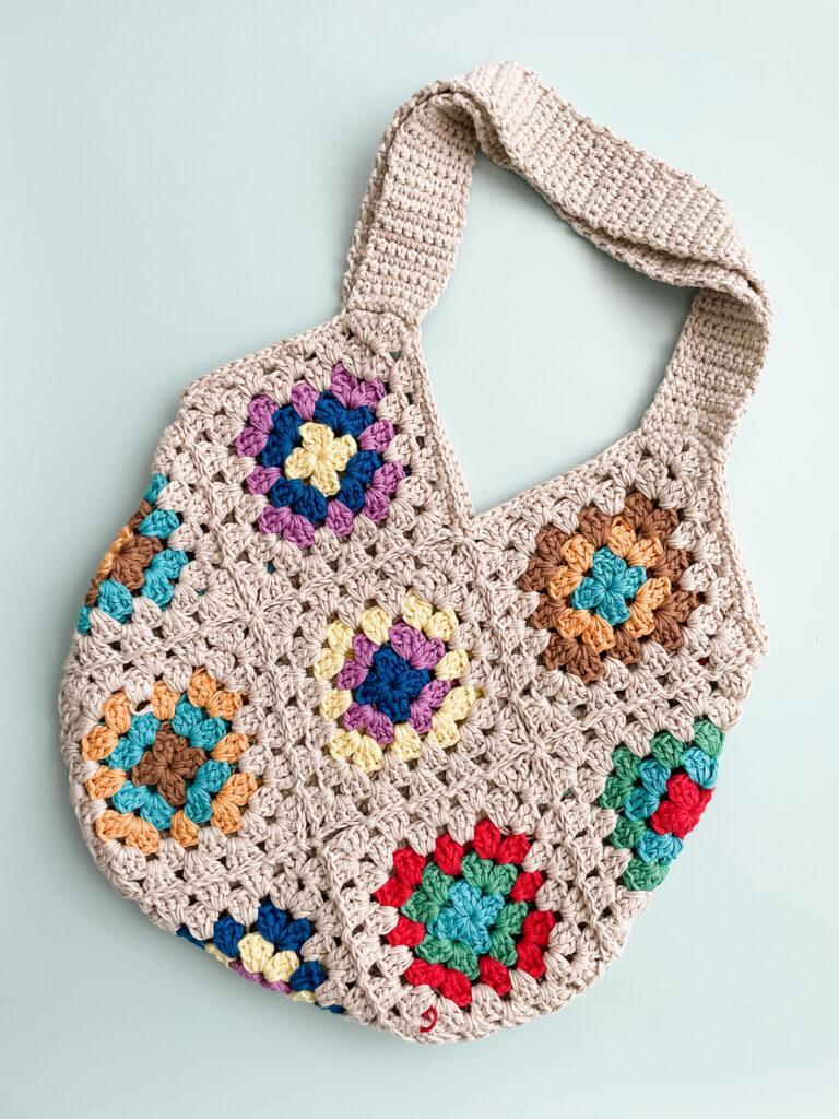 crochet granny square tote bag lay flat on a pale blue background. the main bag colour is beige and the granny squares are made up of alternative colours: yellow, green, blue, red, orange and brown