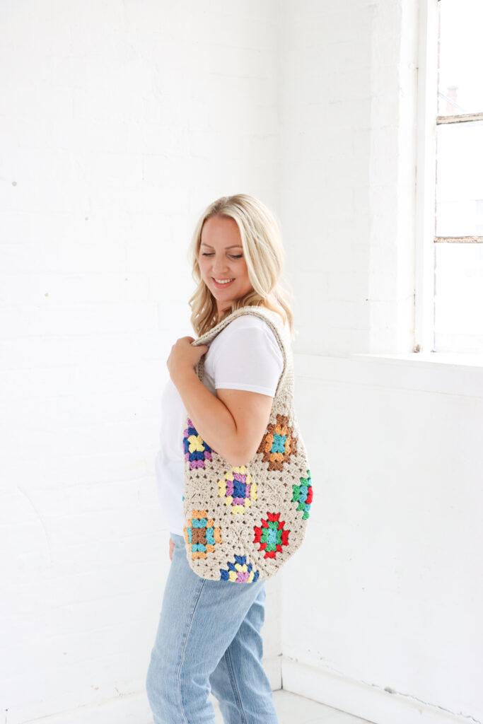 female stood in a white room wearing a white t-shirt and pale blue jeans with a crochet granny square tote bag on her shoulder.