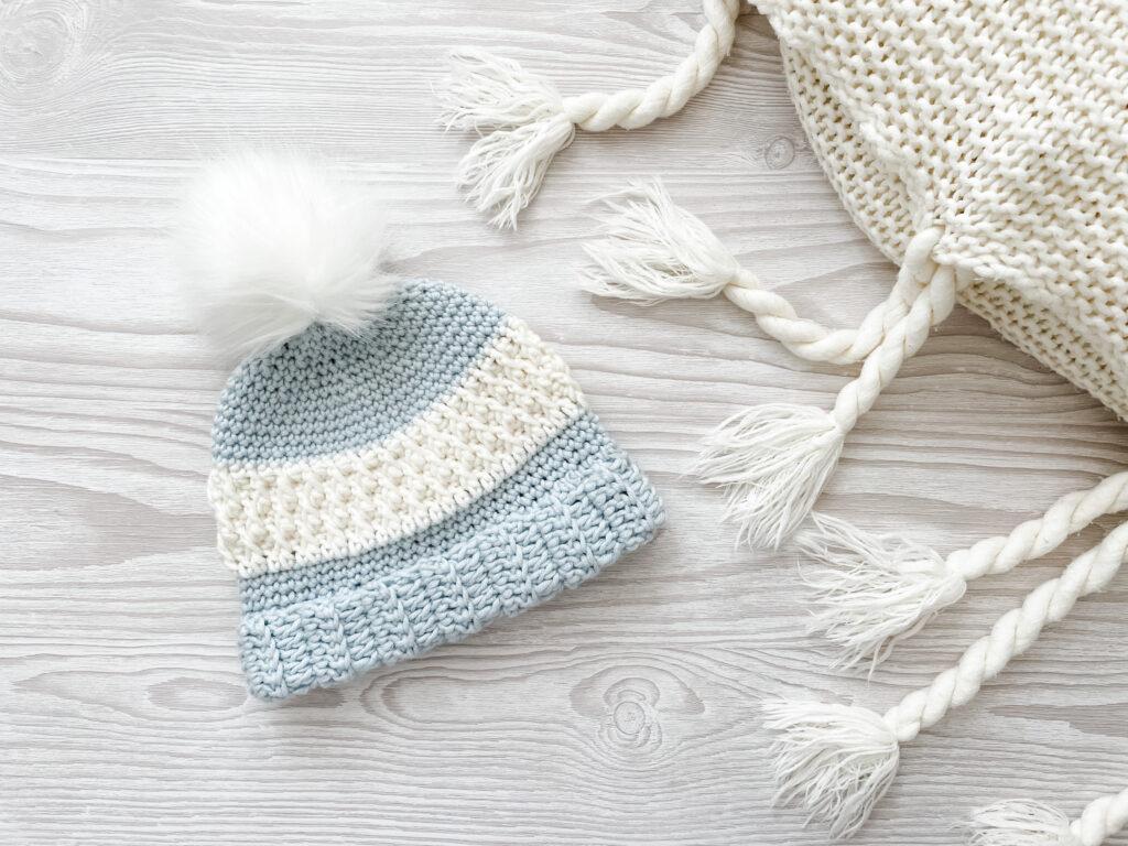 The Alaskan Delights hat sits on a pale grey wooden background. Made from a pale blue and white yarn combination and topped with a white fluffy pompom, the hat is most prominently made using the alpine stitch.