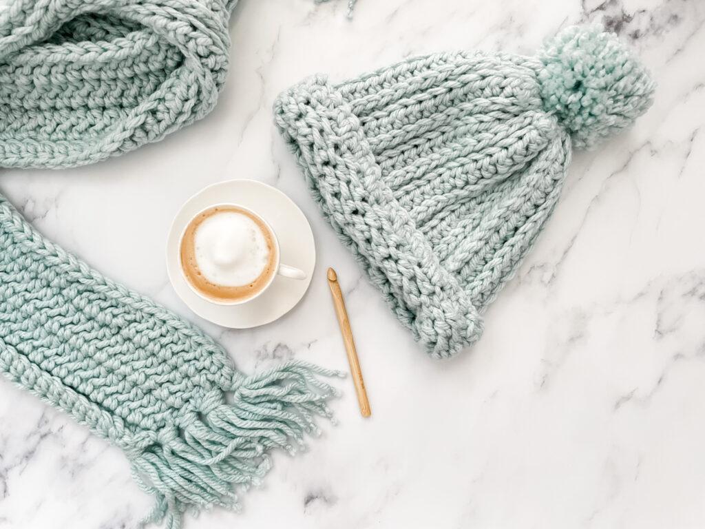 A pale blue hat and scarf set made from chunky yarn are draped over a white marble background. Beside them sit a wooden crochet hook and a frothy coffee.