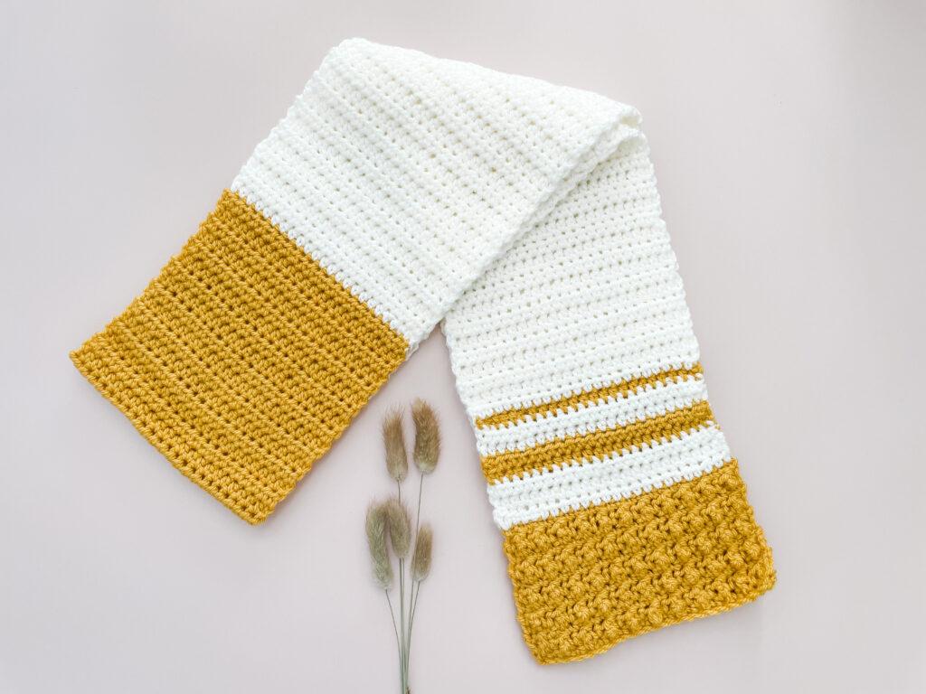 The honey mustard scarf, made in white and mustard yellow yarn sits on a white background beside some natural coloured catkins.