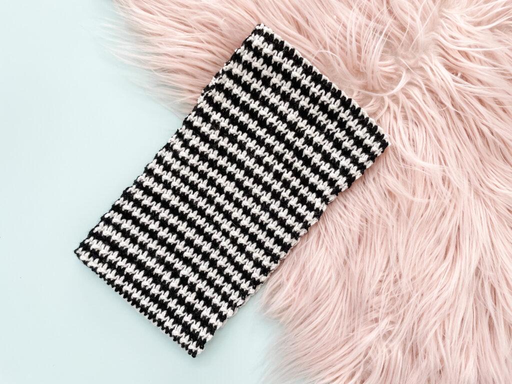 A black and white cowl in the classic houndstooth pattern sits on a pink, furry rug over a white background.