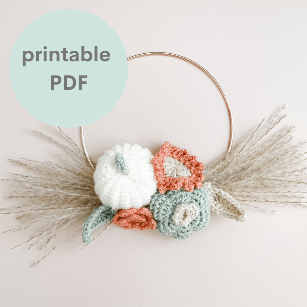 an autumn wreath made onto a gold hoop lay flat on a cream backdrop. The wreath is made up of crochet pumpkins, flowers and leaves with pampas grass at either side of the cluster of crochet flowers, pumpkins and leaves.