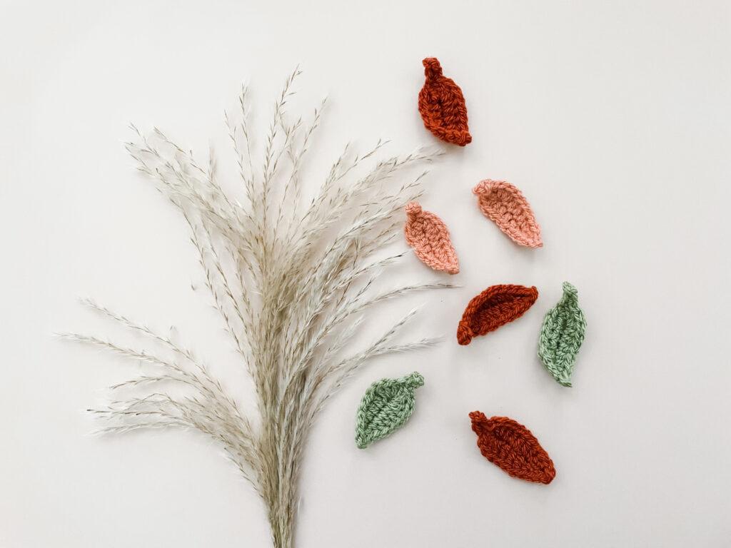 a section of crochet leaves made in rust, orange and green yarn lay flay on a cream backdrop alongside a small bunch of pampers grass.