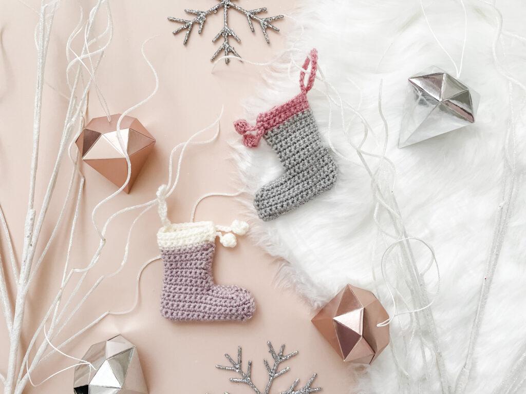two mini crochet stockings lay flat on a nude pink back drop with a white fur rug to the right of the backdrop surrounded by white sparkly branches, diamond-shaped decorations in metallics and two sparkly snowflakes