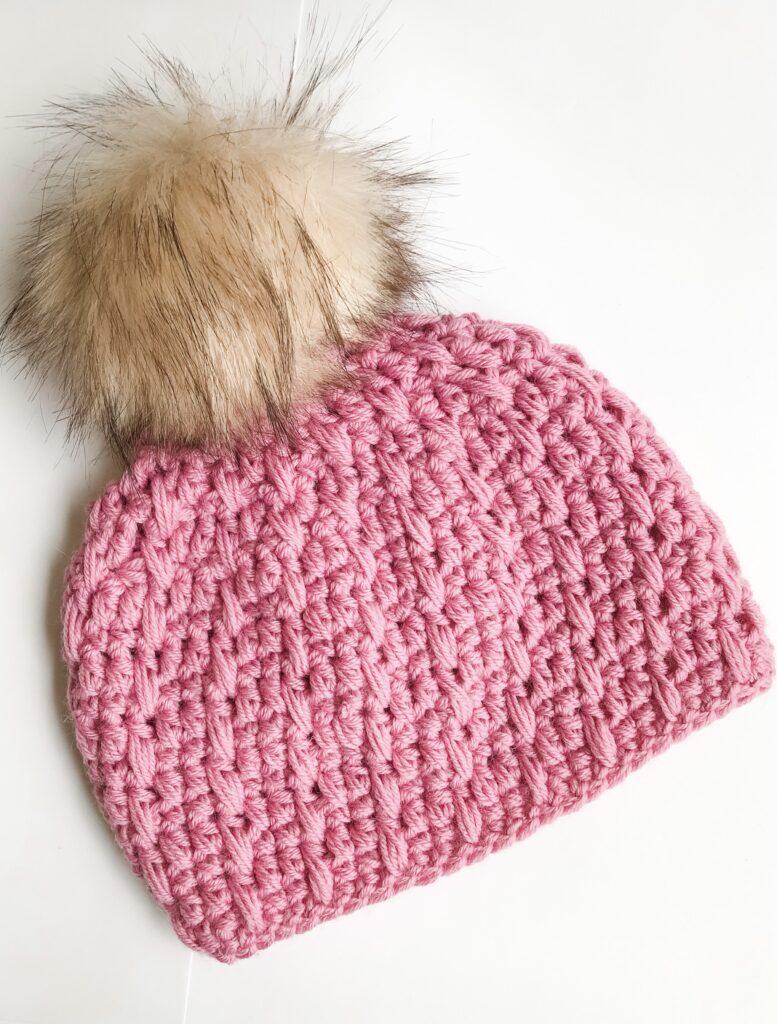 a pink crochet beanie hat with a camel coloured fur pom pom lay flat on a white background