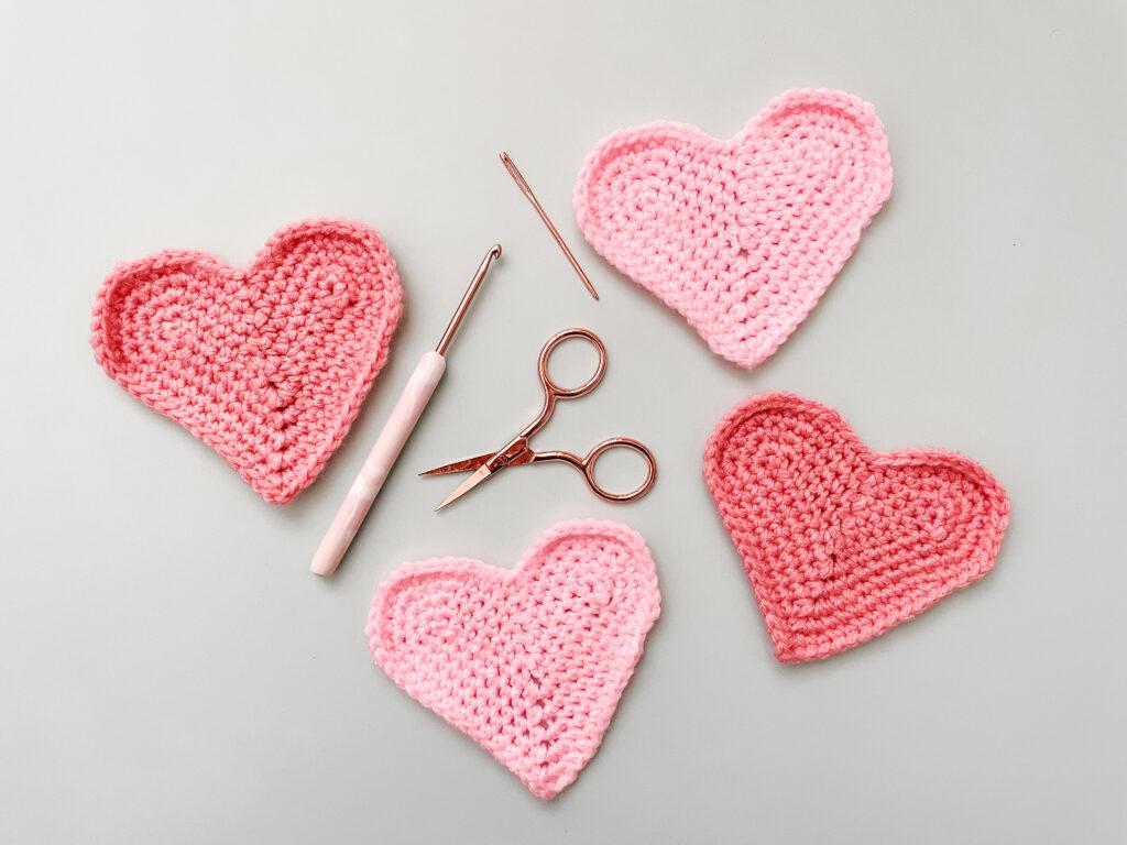flat lay image of four heart coasters on a pale blue background with a rose gold crochet hook, scissors and darning needle.