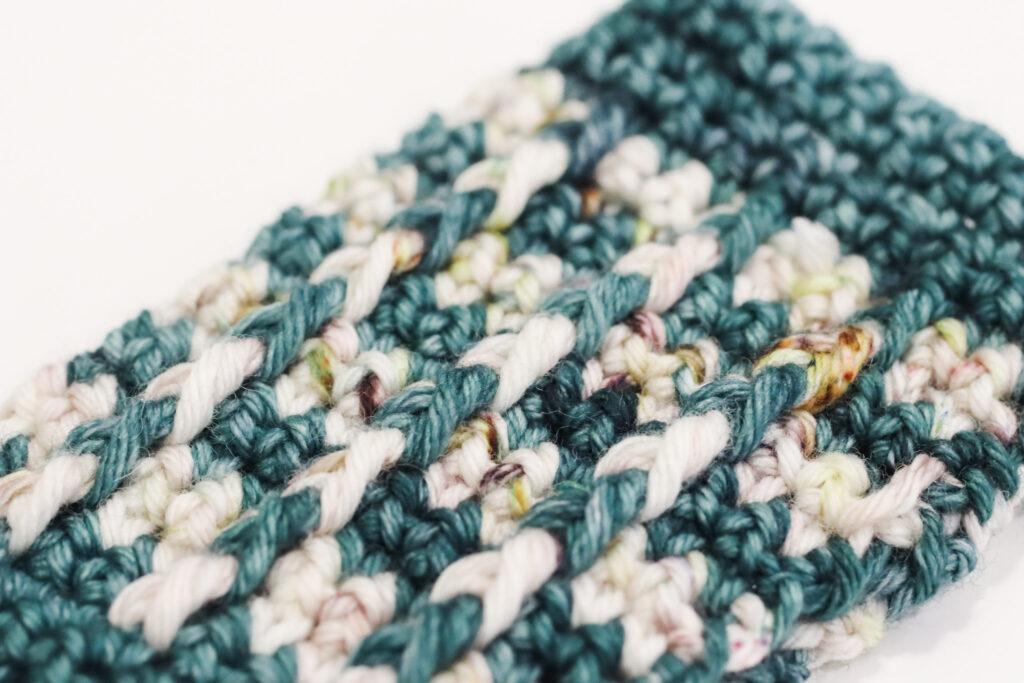 a close-up image of a crocheted wrist warmer in green and cream indie-dyed yarn. The image shows the texture of the bar stitch using indie-dyed yarn.