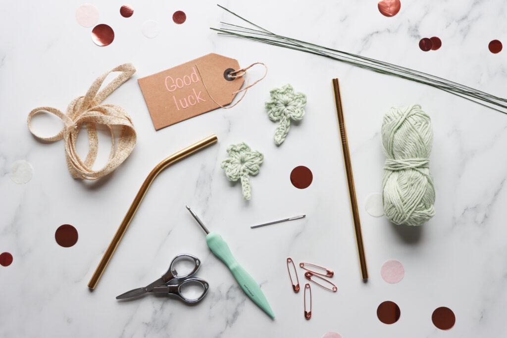 a flat lay of equipment including a metal straw, gold ribbon, crochet hook, scissors, darning needle, pale green yarn, crochet shamrock, safety pins and thin craft wire.