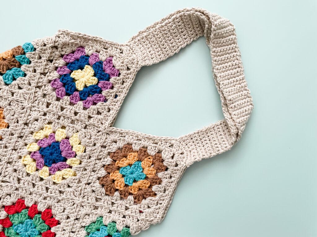 a crochet granny square tote bag lay flay on a pale blue back ground.