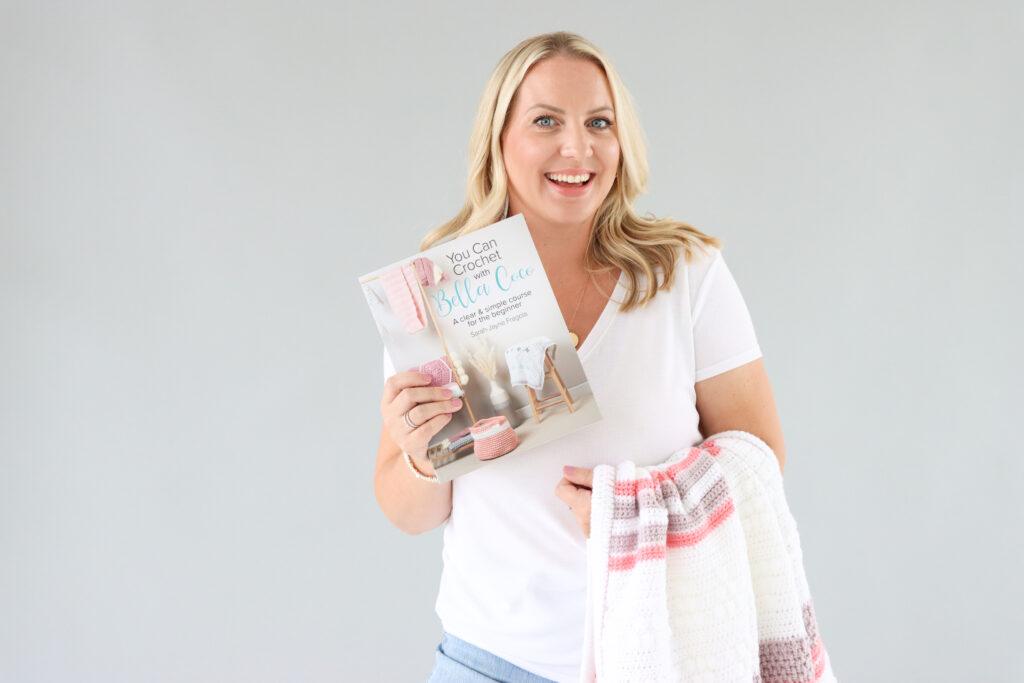 Sarah-Jayne from Bella Coco stood infont of a grey background with a crochet blanket draped over her arm whilst holding her new book in the opposite hand.