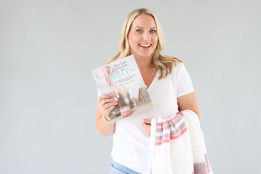 Sarah-Jayne from Bella Coco stood infont of a grey background with a crochet blanket draped over her arm whilst holding her new book in the opposite hand.