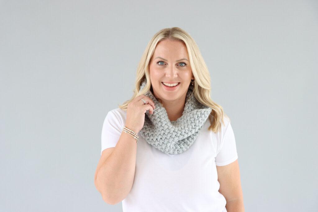 female stood in front of a grey background wearing a white t.shirt and grey crochet cowl.