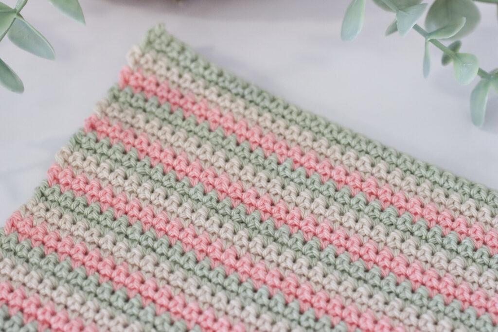 a swatch of UK double crochet using three colours of yarn in paintbox cotton dk in colours: pistachio, vanilla cream and blush pink. The swatch of double crochet is lay on a marble background with a green trailing palm tin the top left corner of the image.