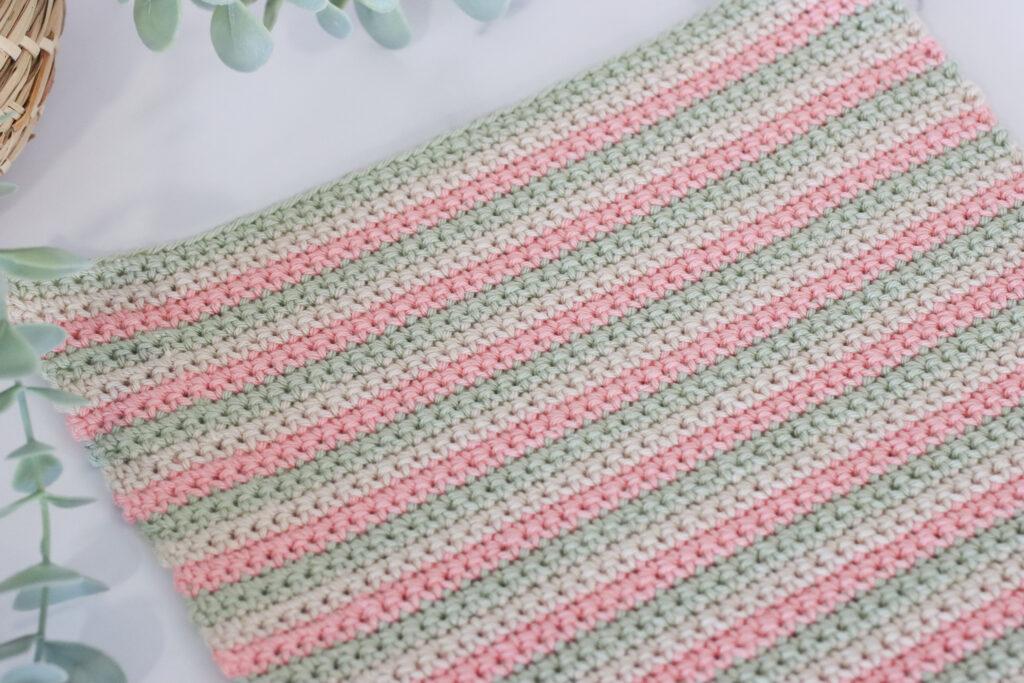 The back facing swatch of UK double crochet using three colours of yarn in paintbox cotton dk in colours: pistachio, vanilla cream and blush pink. The swatch of double crochet is lay on a marble background with a green trailing palm tin the top left corner of the image.