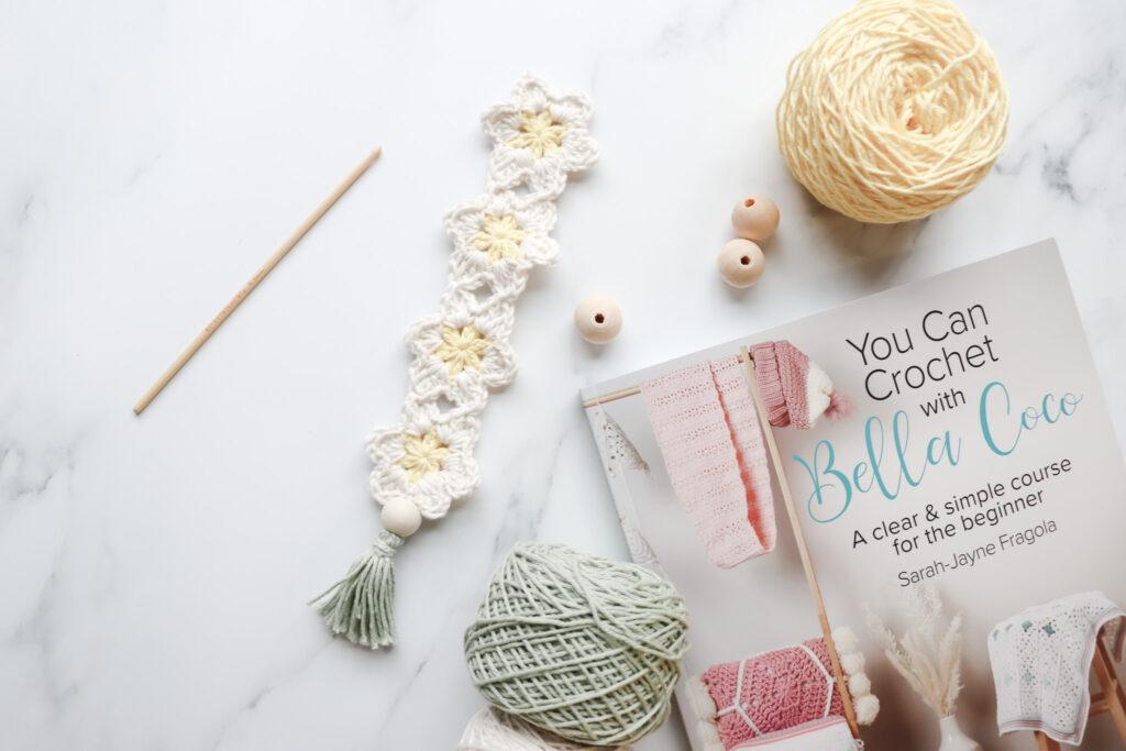 Crochet Daisy Bookmark alongside a wooden crochet hook, 3 wooden beads, a yellow, green and white yarn cake and the Bella Coco Crochet book.