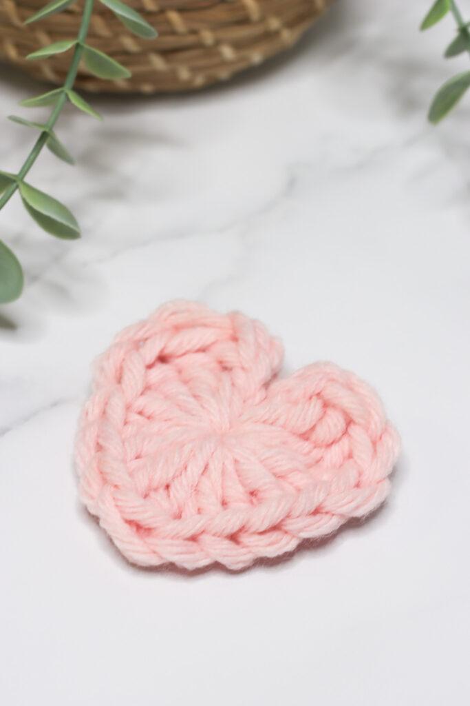 Crochet heart made in light pink chunky yarn laid on a marble background with a hint of green foliage