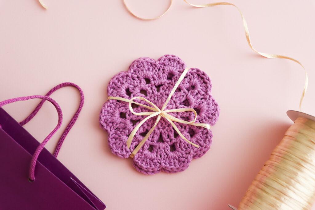 a flat lay image of two purple crochet ocasters tied together with gold ribbon lay on a pale pink backdrop with the bobbin of ribbon to the right of the image and a purple gift bag appearing in the bottom left of the image.