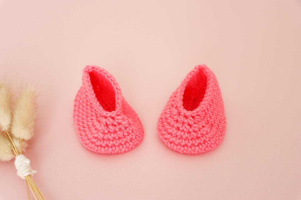 a pair of pink crochet baby booties on a pink background with a small bunch of dried flowers in the left corner of the image.