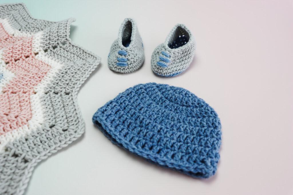 A blue baby hat lay flat on an ombre back drop green going into pink, with a pair of grey and blue baby booties above the hat and a 12 point star blanket showing in the left side of image.
