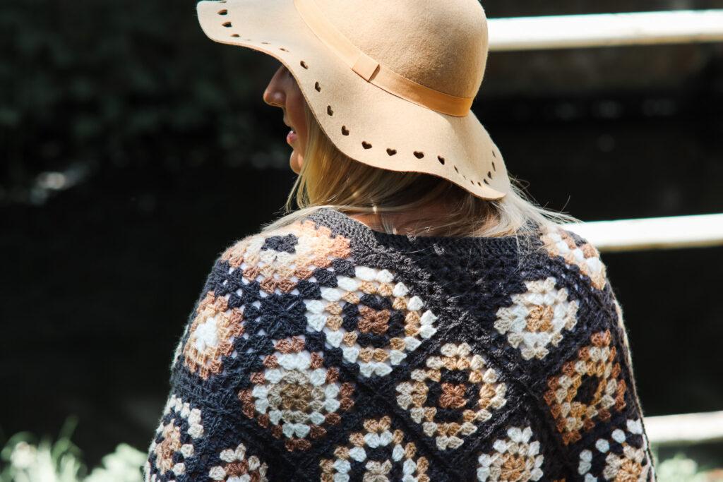 female with should length blonde hair stood in front of a stone bridge wearing a crochet poncho made of granny squares in a variety of colours including: dark grey, cream, camel and brown wearing a camel coloured hat and black skinny jeans.
