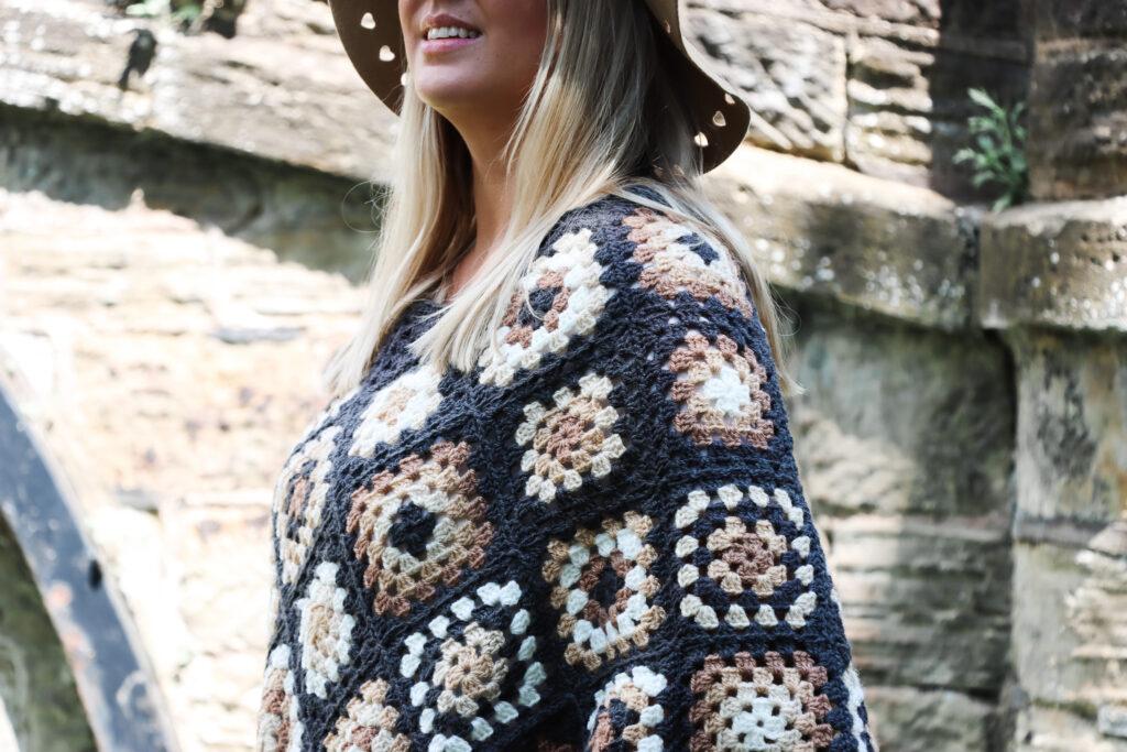 female with should length blonde hair stood in front of a stone bridge wearing a crochet poncho made of granny squares in a variety of colours including: dark grey, cream, camel and brown wearing a camel coloured hat and black skinny jeans.
