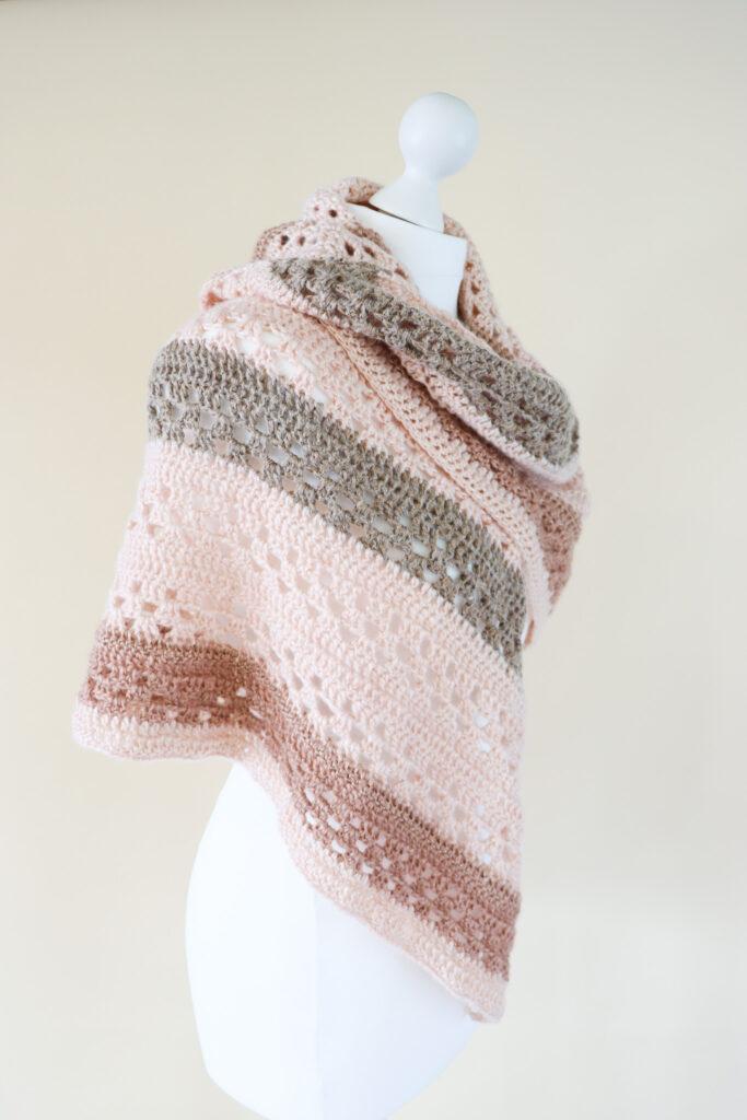 a crochet shawl made is neutrals shades of metallic yarn worn on a white mannequin stood in front of a cream background