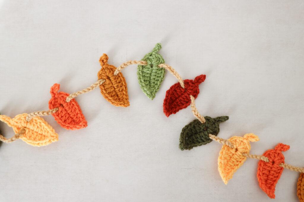 a crochet leaf garland lay flay on pale background. The crochet leaves are made in autumnal shades of green, orange and browns. They are threaded through a crochet chain in a beige yarn.