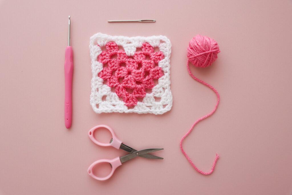a heart granny square with yarn, scissors and a darning needle lay flat alongside on a pink background