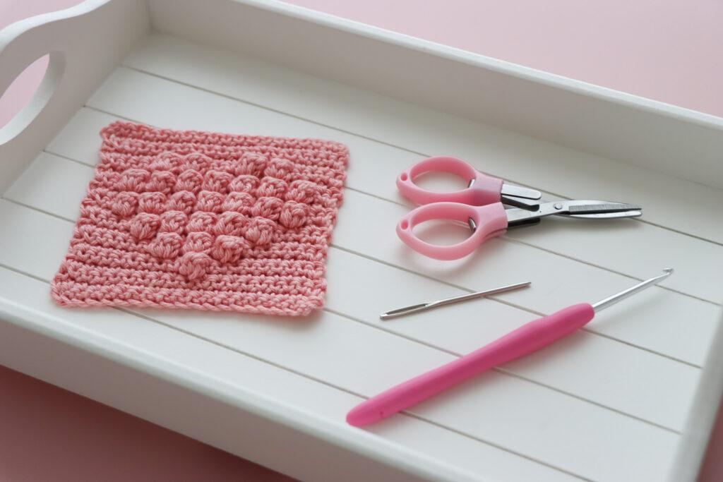 a crochet bobble heart square on a white wooden tray alongside crafting scissors and a darning needle.