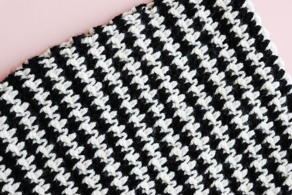 Crochet swatch worked in the houndstooth stitch using black and white yarn, laid on a light pink background and showing a diagonal pattern.